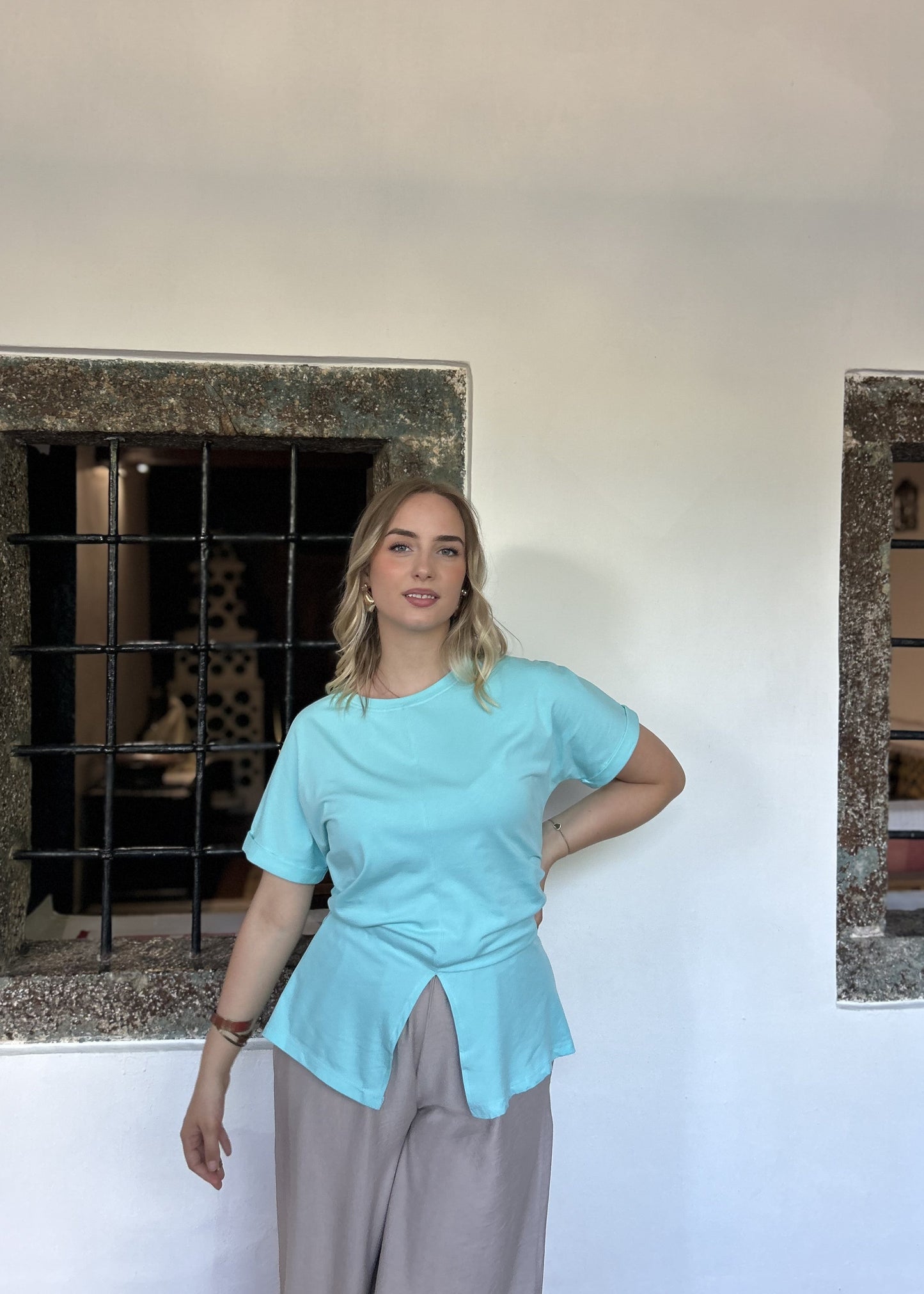 Sindy mint majica uz bježičaste široke pantalone — pogled s rukom na boku ispred kamenih prozora