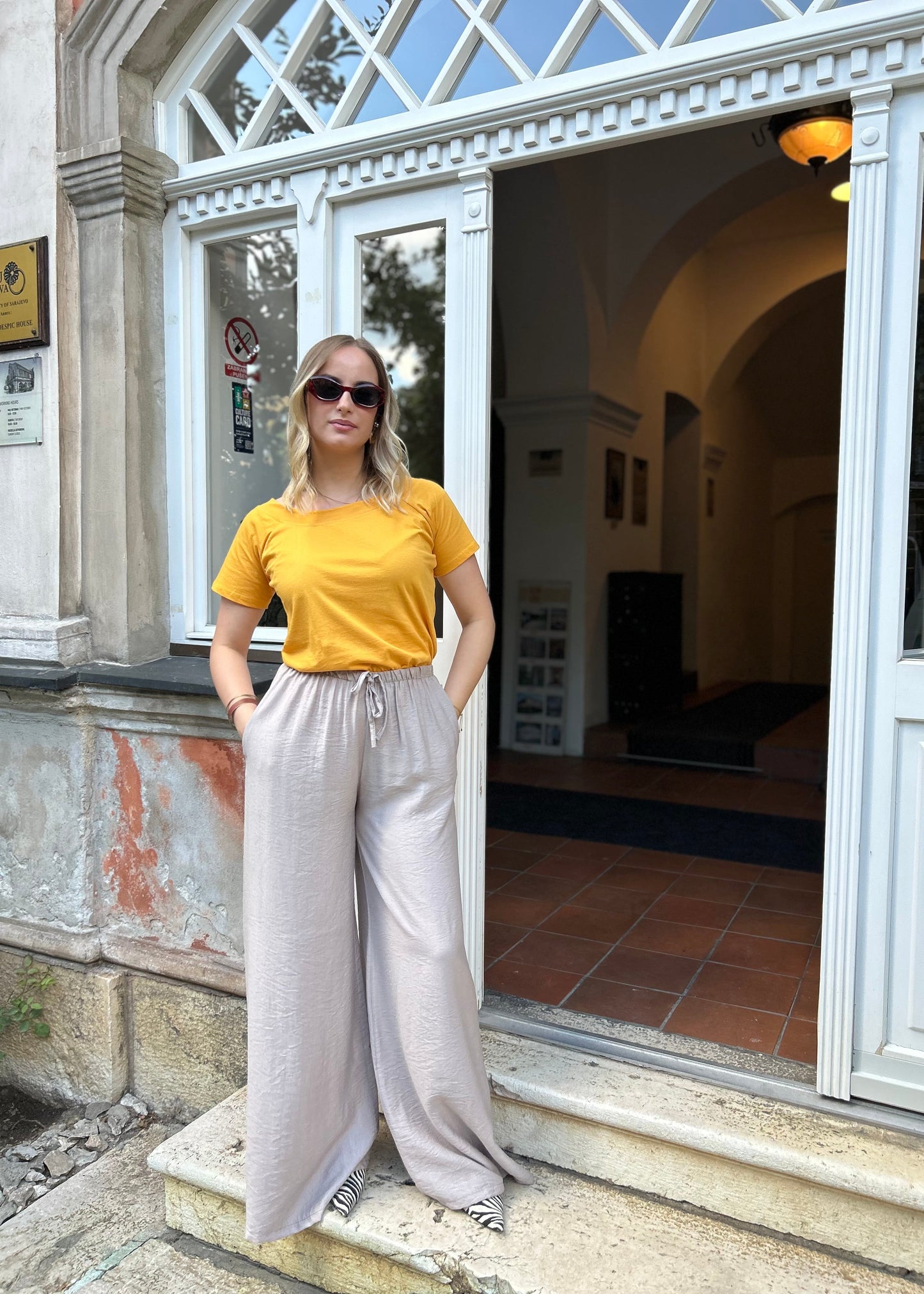Sindy žuta majica uz bježičaste pantalone — puni izgled s rukama u džepovima ispred staklenih vrata