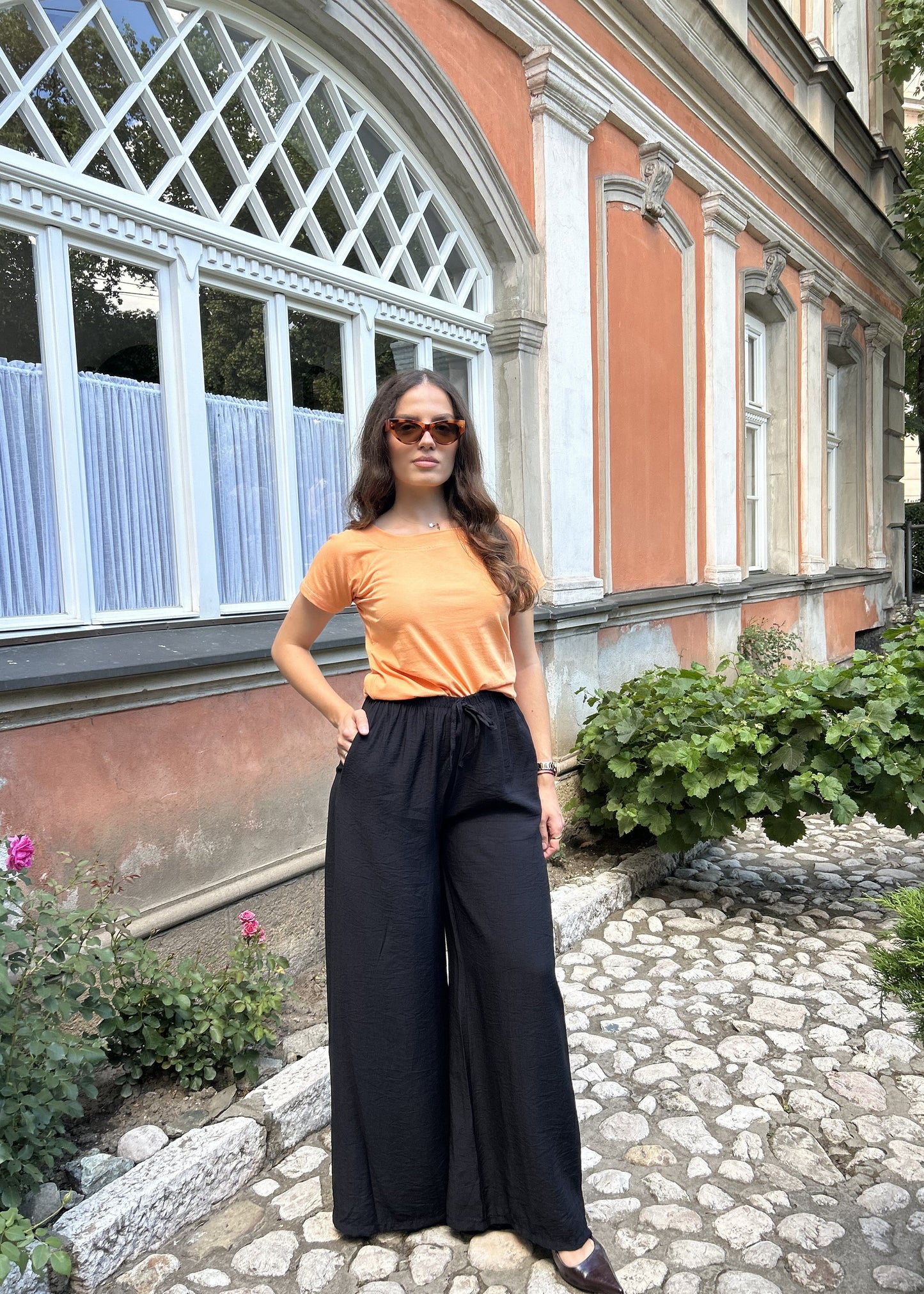 Sindy narandžasta majica — bočni pogled s rukom u džepu crnih pantalona ispred zgrade