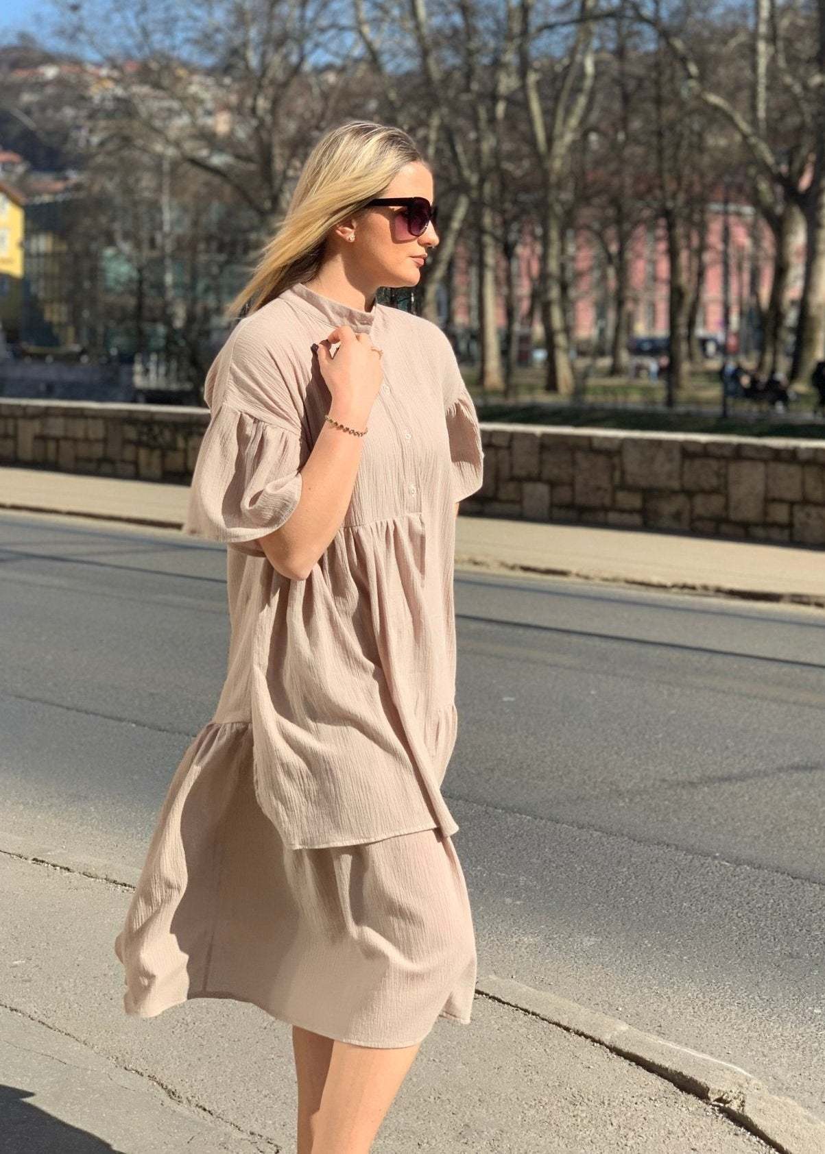 Žena u bež midi haljini Lily s volanima i kratkim rukavima nosi sunčane naočale i crne gležnjače na otvorenom
