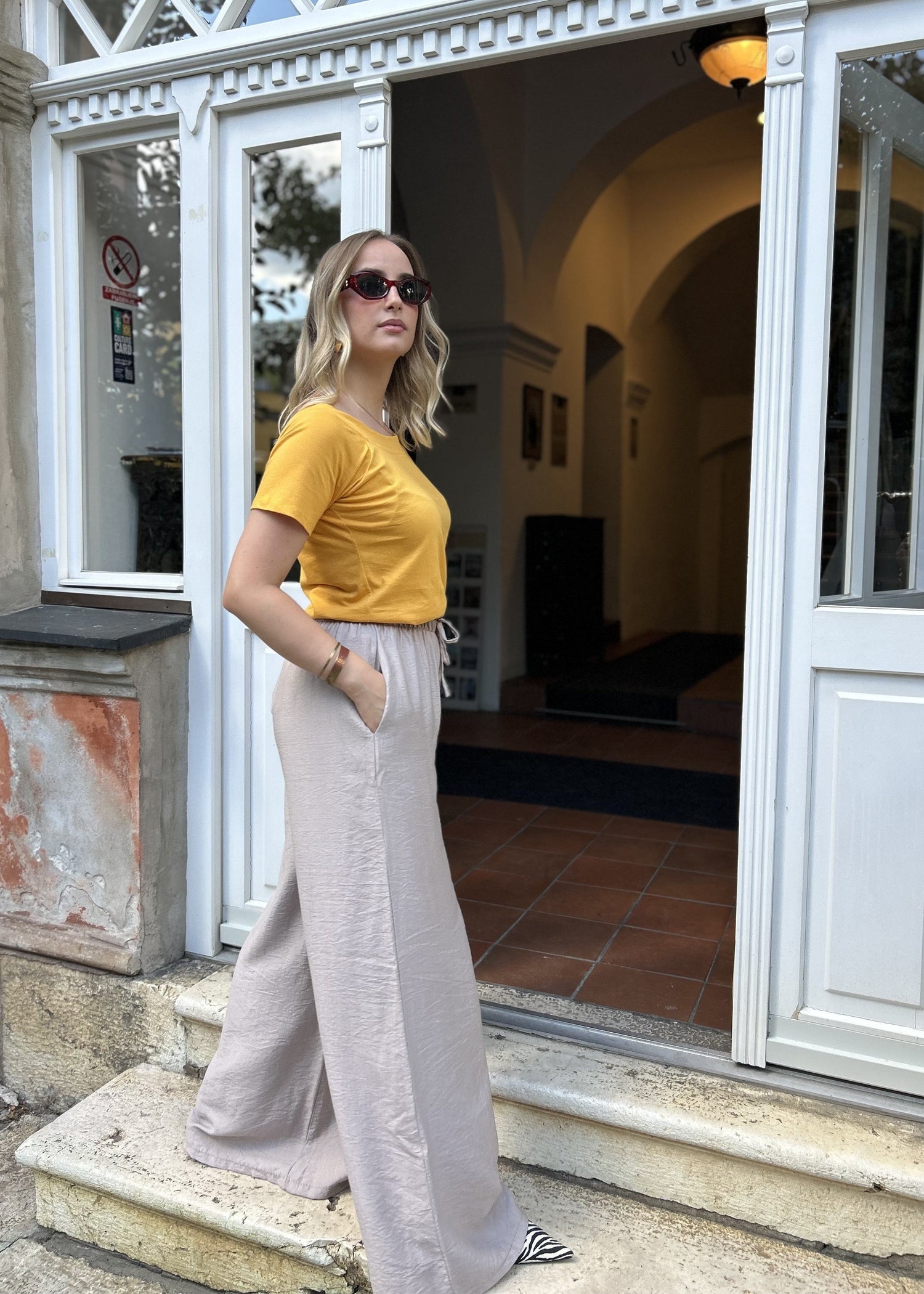 Sindy žuta majica uz bježičaste široke pantalone — bočni pogled na stepenicama historijske zgrade