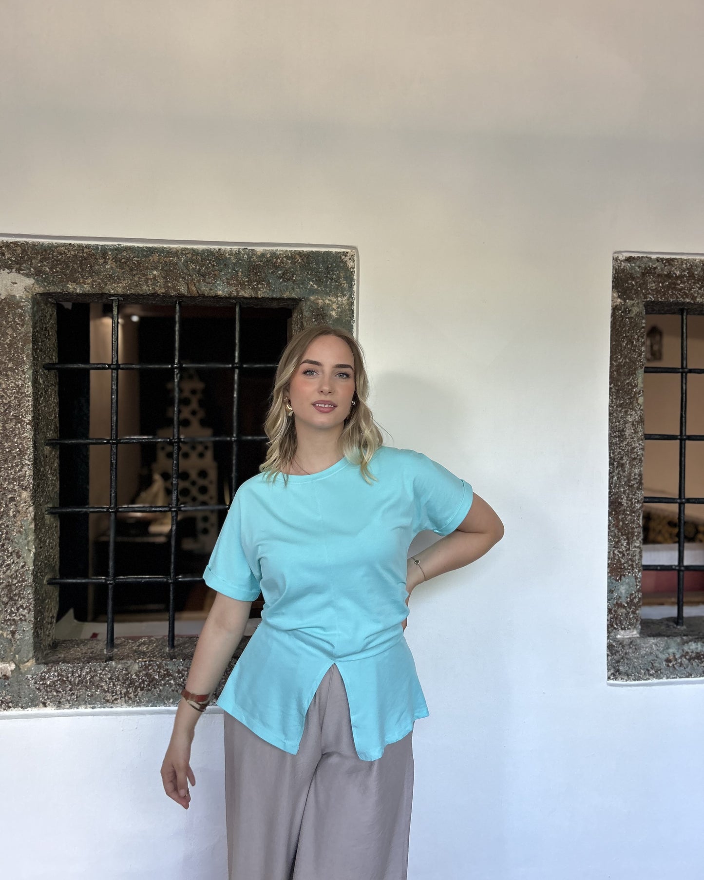 Sindy mint majica uz bježičaste široke pantalone — pogled s rukom na boku ispred kamenih prozora