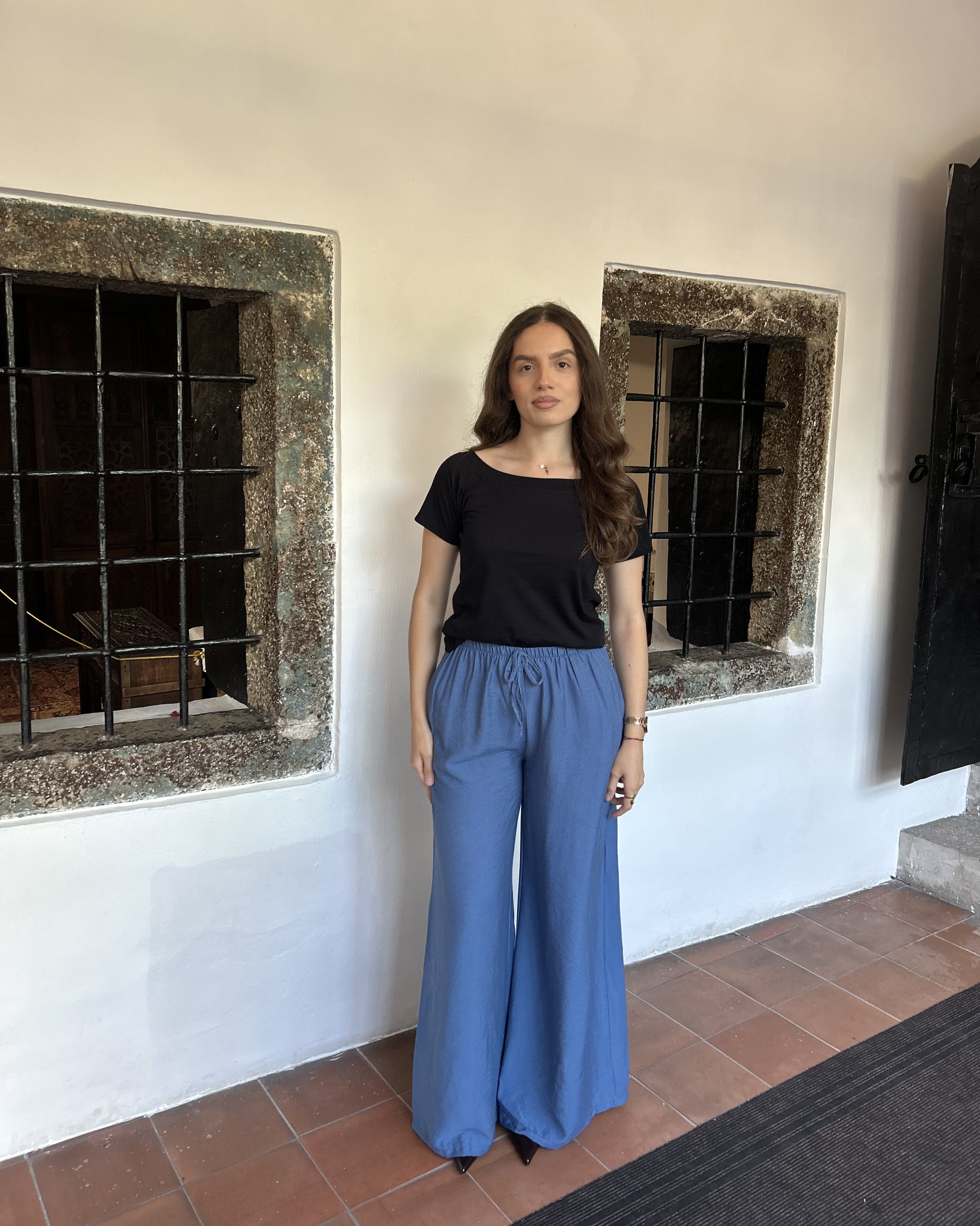 Sindy crna majica s širokim izrezom i plavim širokim pantalonama — puni izgled uz bijeli zid