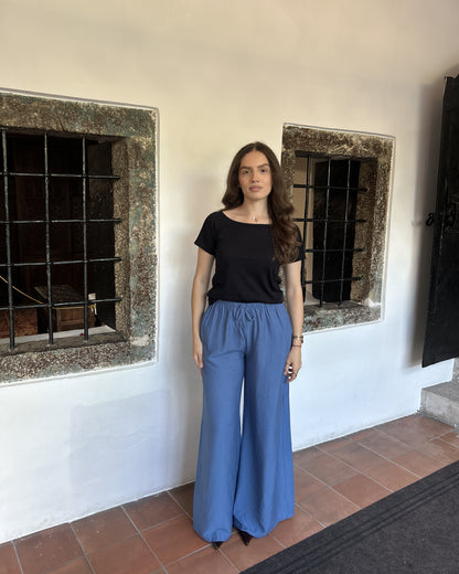 Sindy crna majica s širokim izrezom i plavim širokim pantalonama — puni izgled uz bijeli zid