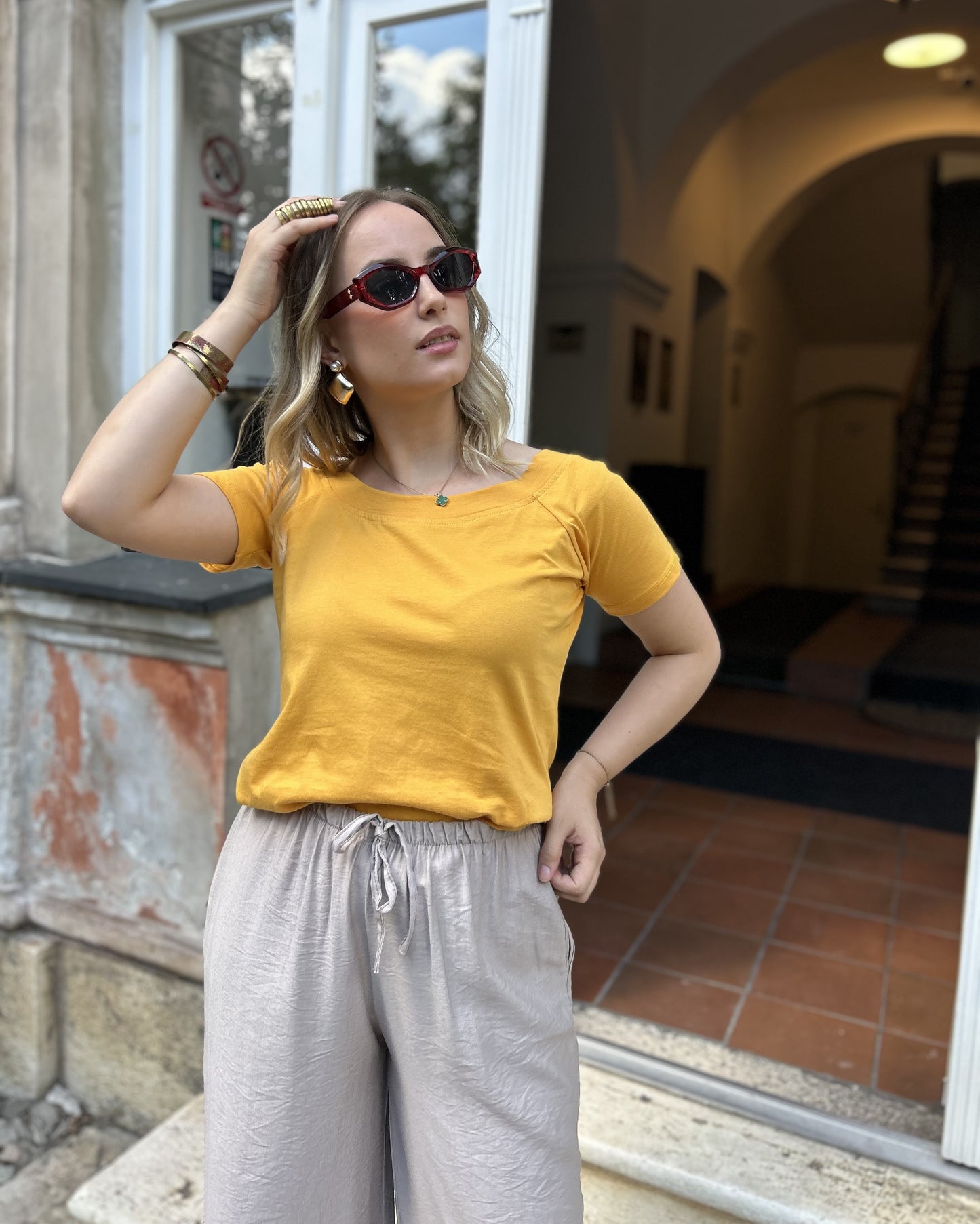 Sindy žuta majica — portretni pogled s zlatnim nakitom i naočalama ispred historijske fasade