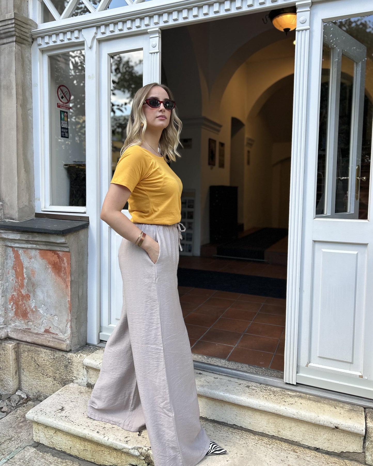Sindy žuta majica uz bježičaste široke pantalone — bočni pogled na stepenicama historijske zgrade