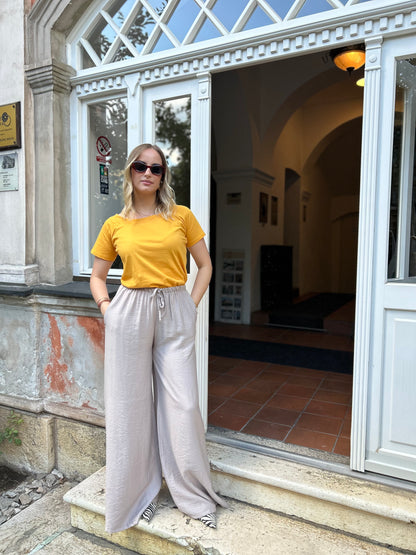 Sindy žuta majica uz bježičaste pantalone — puni izgled s rukama u džepovima ispred staklenih vrata