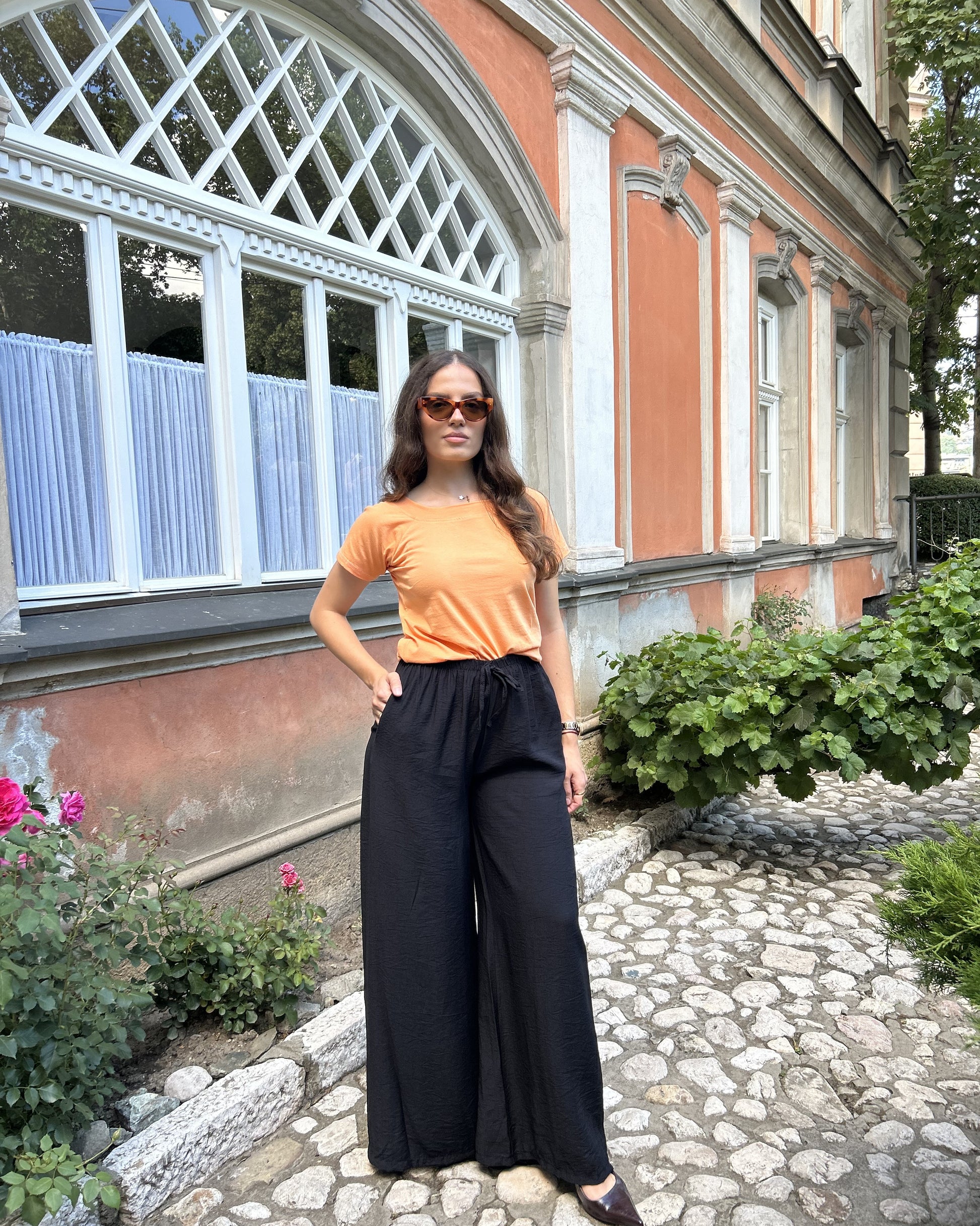 Sindy narandžasta majica — bočni pogled s rukom u džepu crnih pantalona ispred zgrade