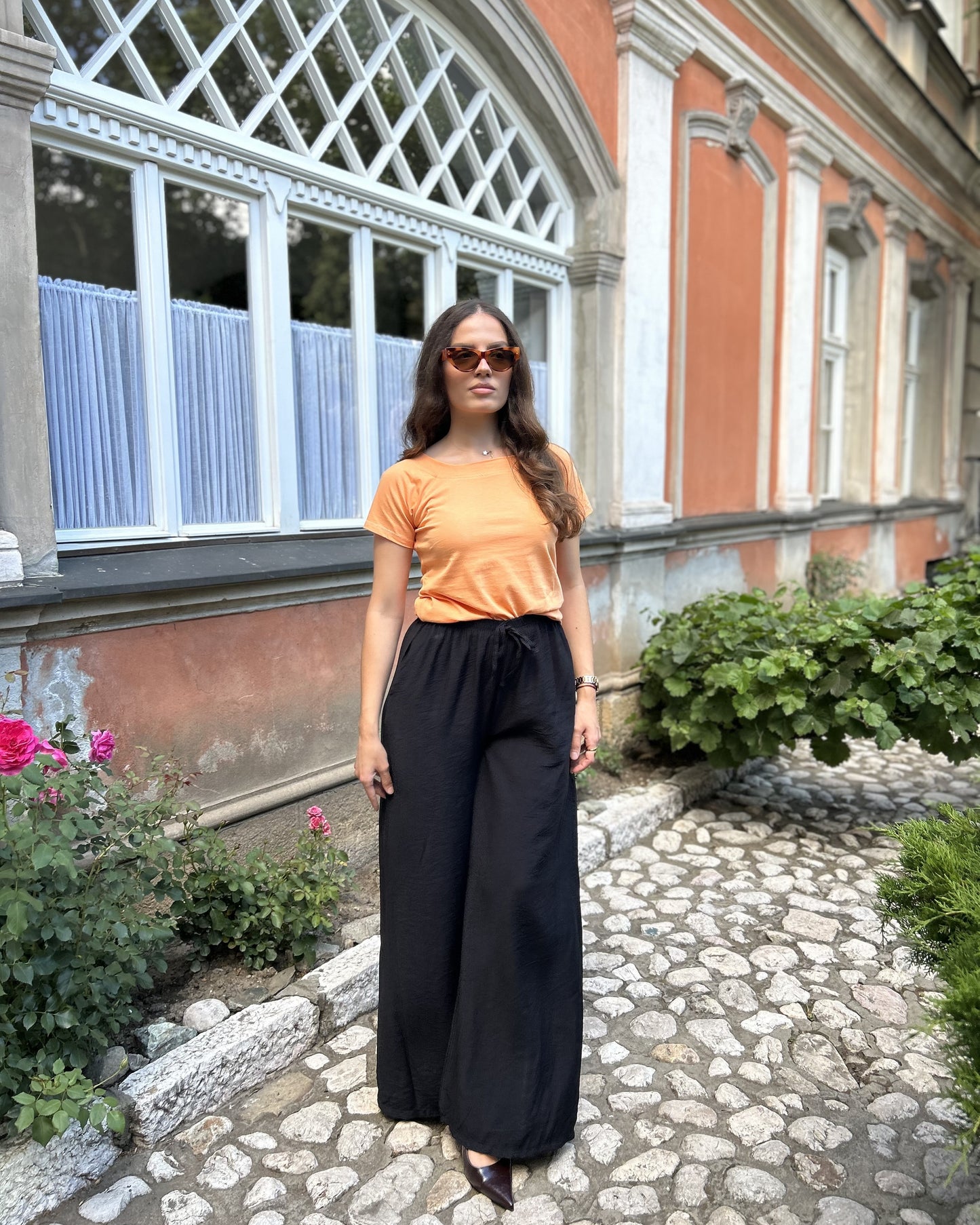 Sindy narandžasta majica uz crne široke pantalone — puni izgled na kaldrmisanoj stazi