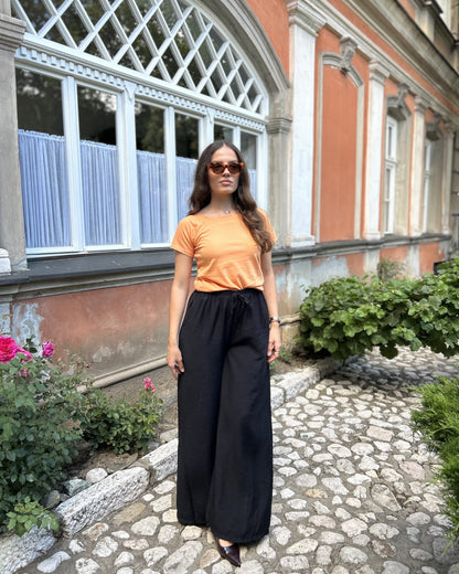 Sindy narandžasta majica uz crne široke pantalone — puni izgled na kaldrmisanoj stazi