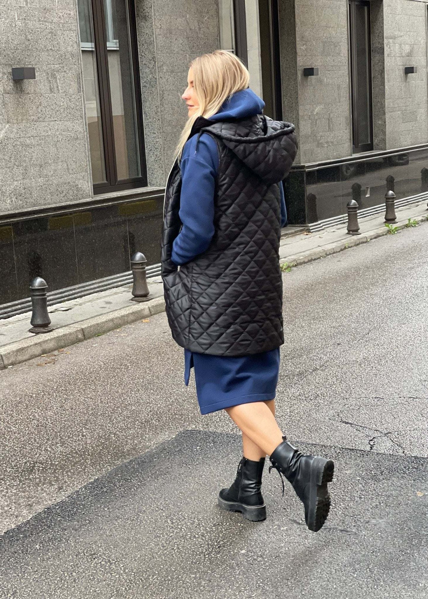 Žena nosi crni prošiveni prsluk s rajsferšlusom preko plave dukserice na otvorenom urbanom okruženju