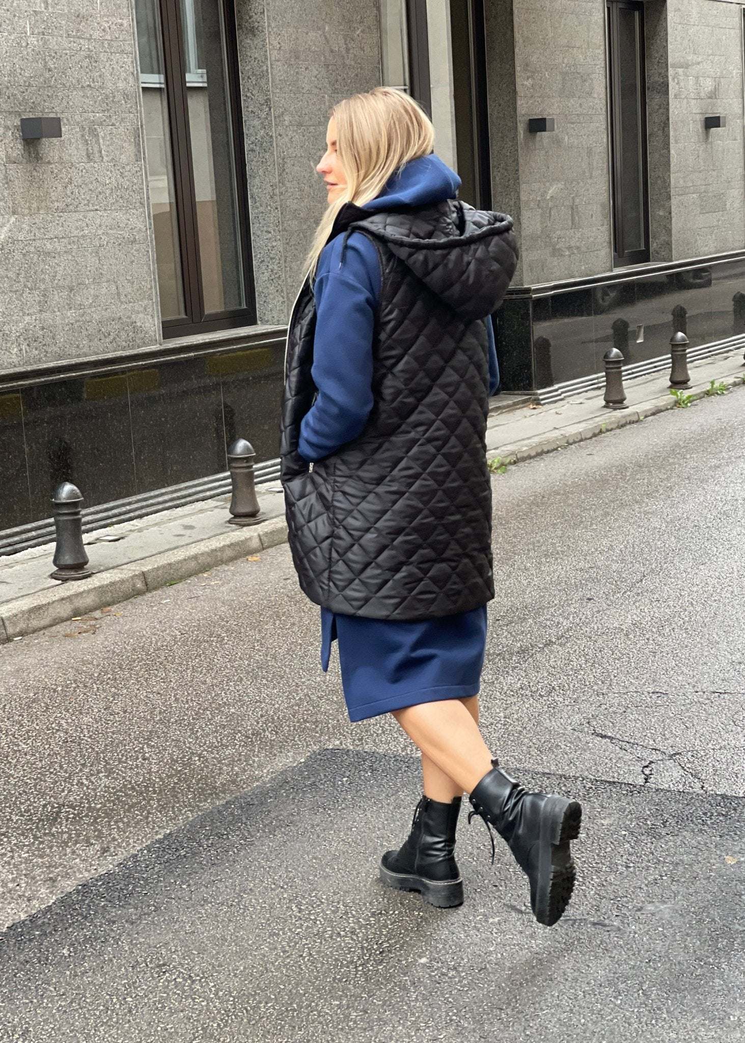 Žena nosi crni prošiveni prsluk s rajsferšlusom preko plave dukserice na otvorenom urbanom okruženju