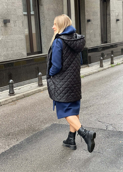 Žena nosi crni prošiveni prsluk s rajsferšlusom preko plave dukserice na otvorenom urbanom okruženju