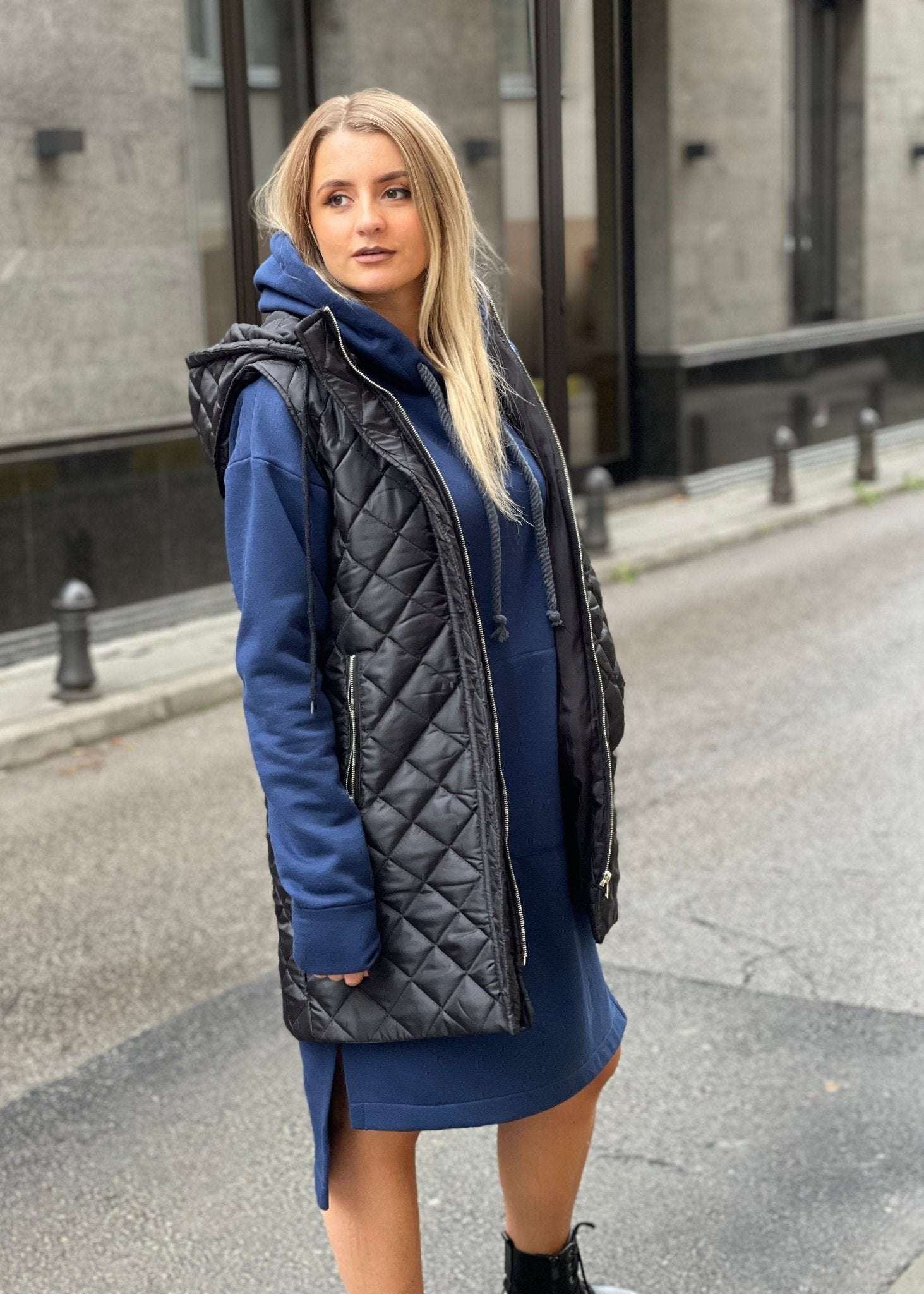 Žena nosi crni prošiveni prsluk s rajsferšlusom preko plave dukserice na otvorenom urbanom okruženju