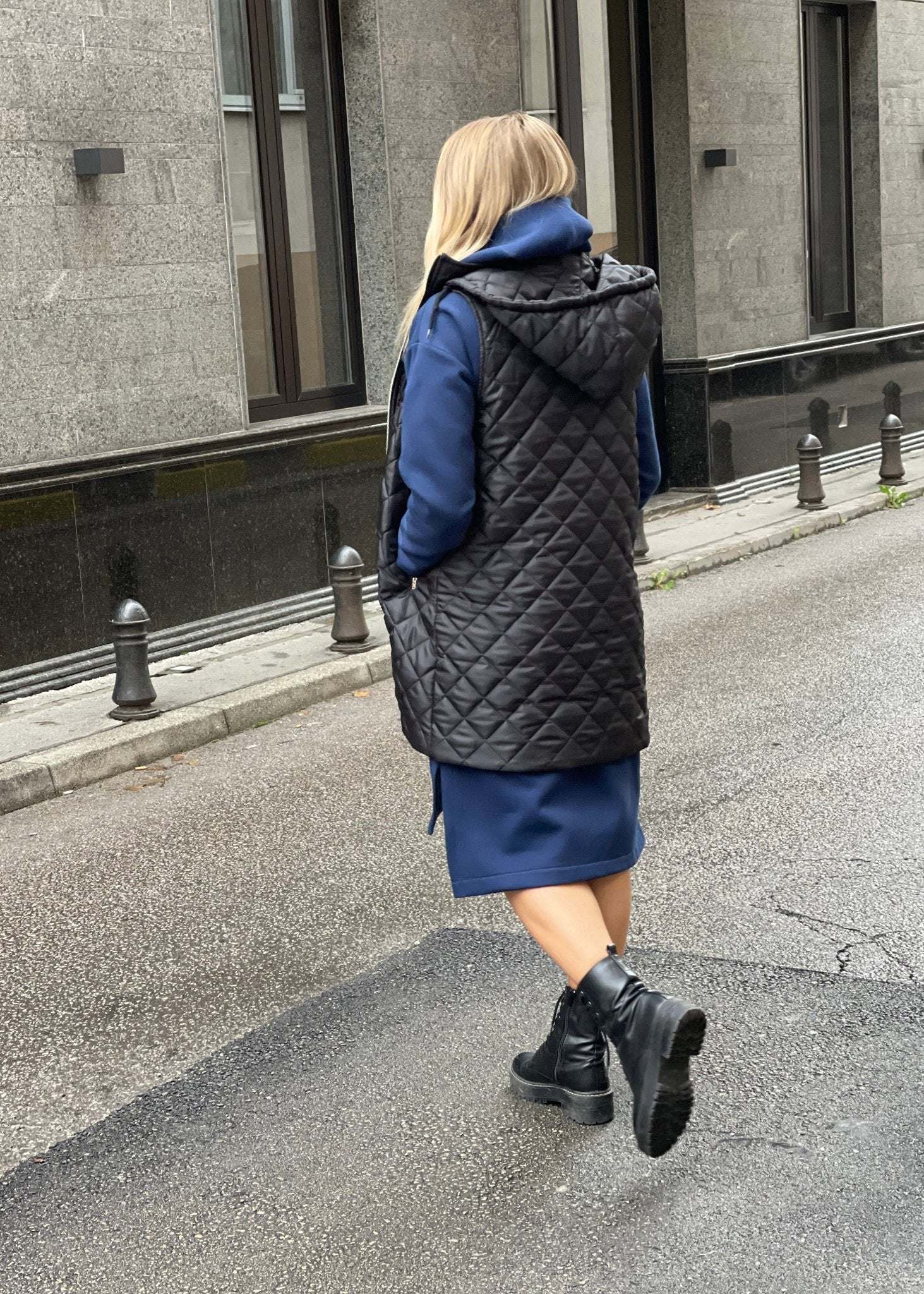 Žena nosi crni prošiveni prsluk s rajsferšlusom preko plave dukserice na otvorenom urbanom okruženju