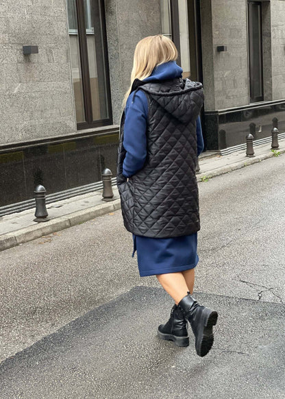 Žena nosi crni prošiveni prsluk s rajsferšlusom preko plave dukserice na otvorenom urbanom okruženju