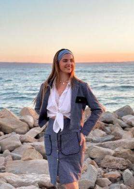 Woman wearing grey-blue tweed komplet with black pocket details standing on rocky shore at sunset