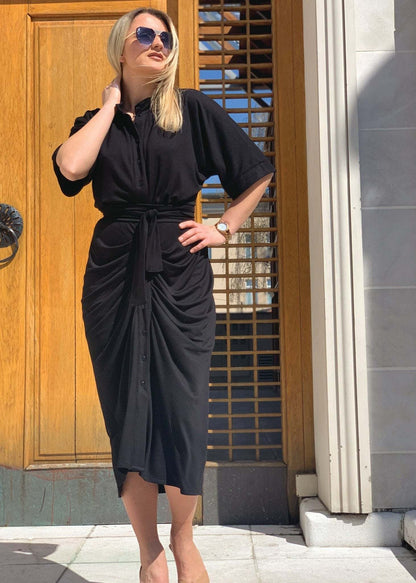 Woman wearing black midi Haljina Nora dress with front pleats and tied waist belt standing outdoors