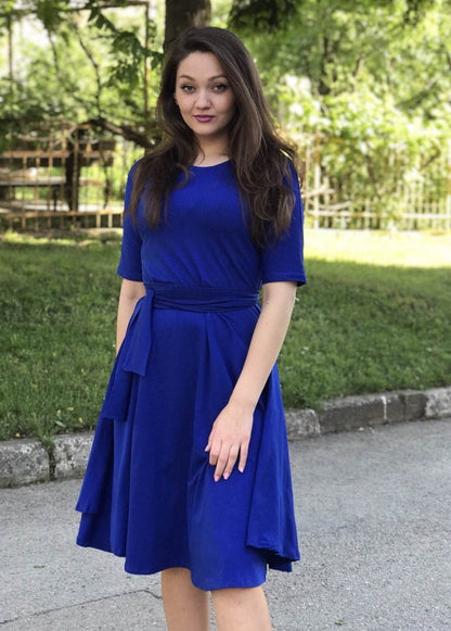Žena u rozoj Haljini Glok s lepršavom midi suknjom i pojasom, stoji na otvorenom