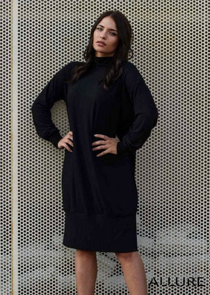 Woman wearing black turtleneck midi dress with long sleeves and slim fit standing against perforated metal background